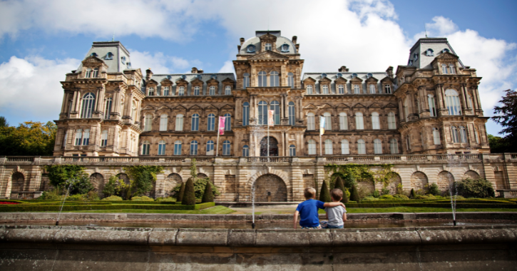 Photograph of a large, ornate historic building with classical architectural features including multiple windows, arches, and decorative stonework under a partly cloudy sky. Two children sit on a low wall in front of a garden with trimmed bushes and fountains, highlighting the building's grandeur and landscaped surroundings.